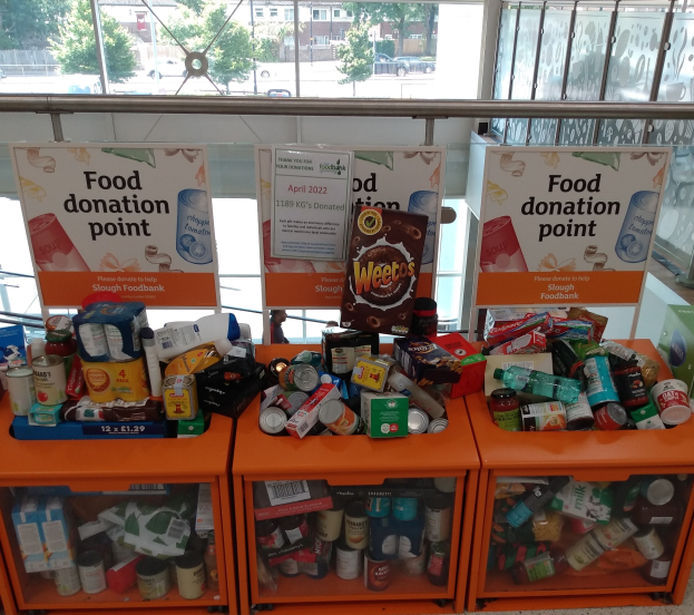 Eine Spendenstelle für Lebensmittel mit orangefarbenen Schäubern, die Flaschen, Dosen, Schachteln und andere Gegenstände enthalten, neben Texttafeln, durch Glasfenster betrachtet, die Fahrzeuge, Bäume, Gebäude und Himmel zeigen.