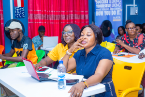 Eine Gruppe von Menschen sitzt um einen Tisch mit Laptops, Flaschen, Mobiltelefonen, Papieren, Stiften und anderen Gegenständen, mit einer Wand mit einem Vorhang und einer Fahne mit Text sowie Flaggen im Hintergrund, die wahrscheinlich an der 2019 Maya-Führungs конференz teilnehmen.