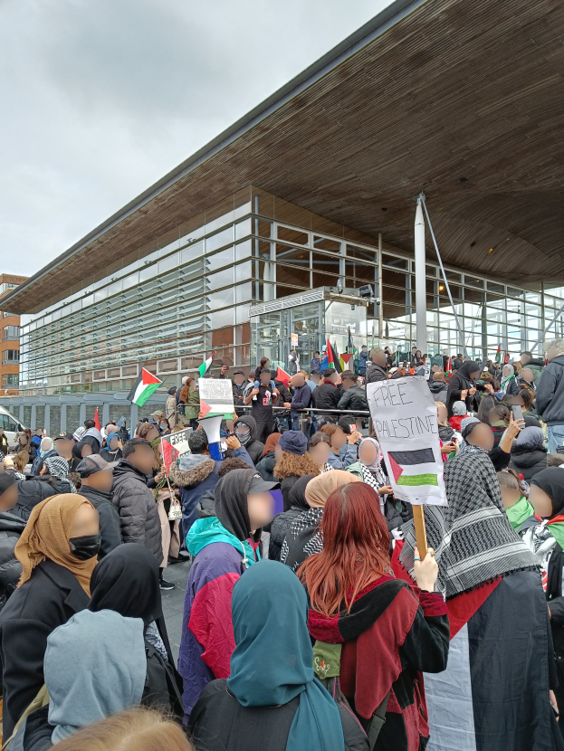 Eine große Gruppe von Menschen steht vor einem Gebäude und hält Schilder und Fahnen in den Händen, während ein Fahrzeug auf der linken Seite und der Himmel oben im Bild zu sehen ist.
