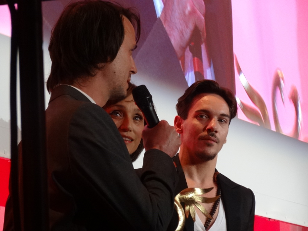 Eine Gruppe von Menschen in Mänteln steht zusammen, einer hält ein Mikrofon und eine Auszeichnung, mit einem Podium und einem Bildschirm im Hintergrund.