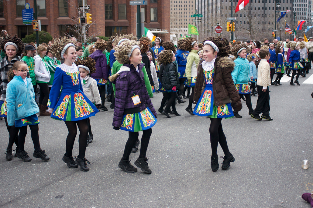 Eine Gruppe von M√§dchen in K√∂rpern mit Jacken und Pullis, die auf einer Stra√Ÿe tanzen, mit einem roten Geb√§ude und einem gelben Verkehrszeichen dahinter und Geb√§uden und Flaggen an Stromm√§sten im Hintergrund.