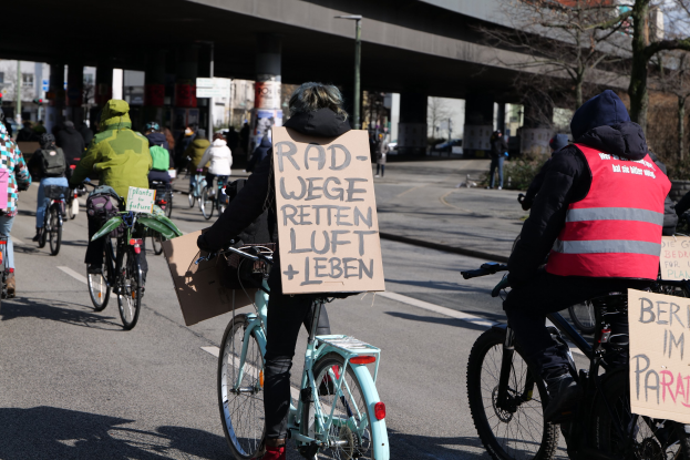Eine Gruppe von Menschen, die auf Fahrrädern eine Straße entlangfährt, mit einer Brücke im Hintergrund, Bäumen an den Seiten und Gebäuden in der Ferne, die Schilder mit verschiedenen Botschaften halten.