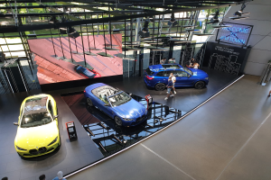Eine Gruppe von Menschen im BMW Museum in München, Deutschland, mit Autos zur Linken und einem Schild mit der Aufschrift "BMW Museum München" zur Rechten, überblickend eine überdachte Stadtlandschaft durch Glasfenster.