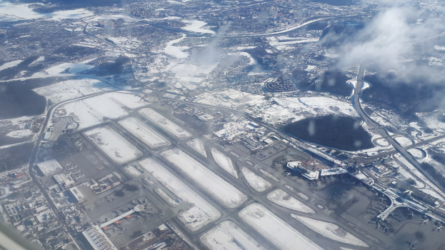 Eine Luftaufnahme eines Flughafens mit schneebedeckter Erde, Gebäuden, Straßen, Brücken, Wasser, Bäumen und Wolken am Himmel.