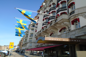 Eine belebte Stadtstraße mit Fahrzeugen, Fußgängern und Gebäuden, mit schwedischen Flaggen im Hintergrund.