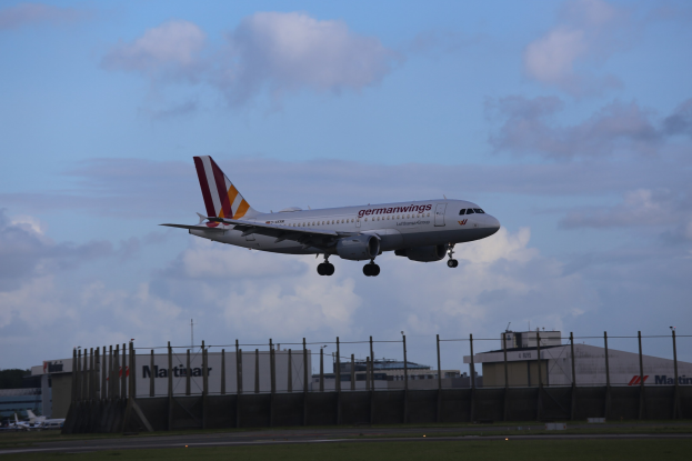 Germanwings Airbus A320-200 beim Abheben vom Frankfurt Airport mit Gras darunter, einem Zaun im Vordergrund, Gebäuden und Bäumen im Hintergrund und einer bewölkten Himmel über.