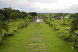 Ein üppiger grüner Park mit einem zentralen Brunnen, umgeben von Bäumen, Pflanzen, Gras, Straßenmasten, Straßenlaternen, Bänken und Menschen unter einem bewölkten Himmel.