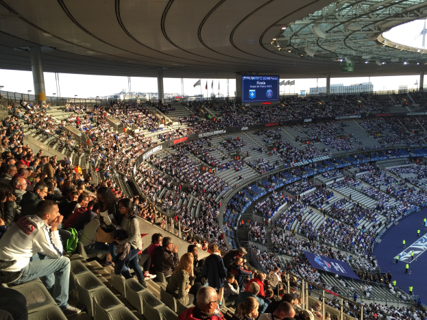Große Zuschauermenge in einem Stadion bei einem Fußballspiel, mit einer Bühne, Fahnen, Stangen, einem Bildschirm und dem Allianz Stadion in München, Deutschland im Hintergrund.