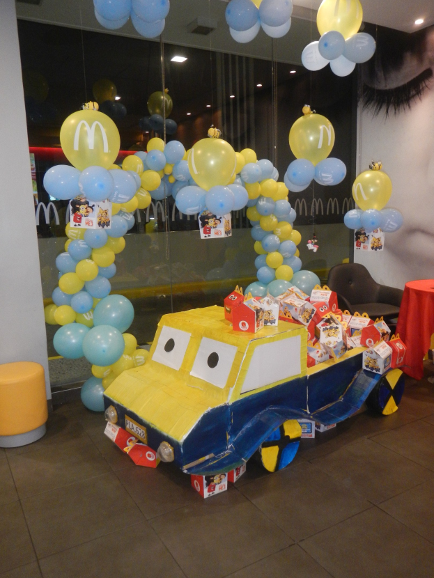Eine McDonald's-Geburtstagsfeier mit einem gelben und blauen Lastwagen, der mit Ballons und anderen Dekoartikeln geschmückt ist, einem rot gedeckten Tisch mit Gegenständen, Hockern und einer Glaswand im Hintergrund und festlicher Deckenbeleuchtung.