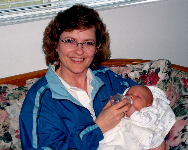 Eine Frau in einer blauen Jacke sitzt auf einem Sofa und füttert ein Baby in der Nähe eines Fensters mit Jalousien.