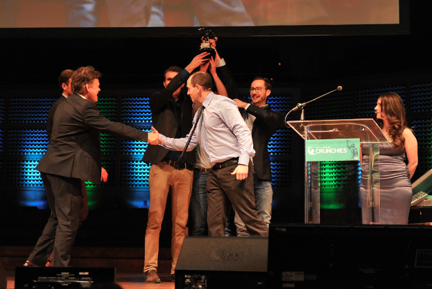 Zwei Männer in schwarzen Mänteln heben einen Preis hoch, lächeln, während ein Mann links die Hand schüttelt und eine Frau rechts steht.