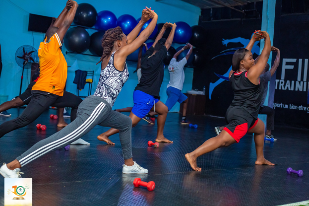 Gruppe von Menschen, die Yoga in einem Fitnessstudio mit Hanteln auf dem Boden machen, ein Banner mit der Aufschrift "Fitness-Training in Ghana" im Hintergrund und diverse Gegenstände wie Ballons, ein Ventilator und ein Stuhl.