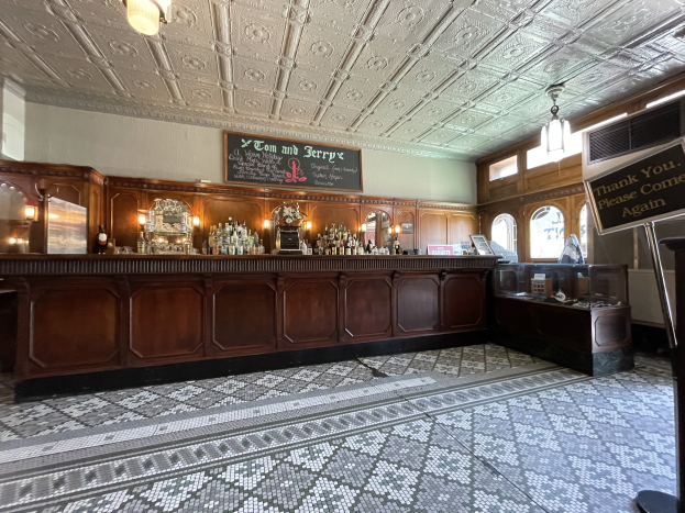 Ein großer Raum mit einer zentralen Bar, Deckenleuchten, Flaschen auf einem Tisch und einer Tafel mit Text auf der rechten Seite, im Tom and Jerry Pub in London.