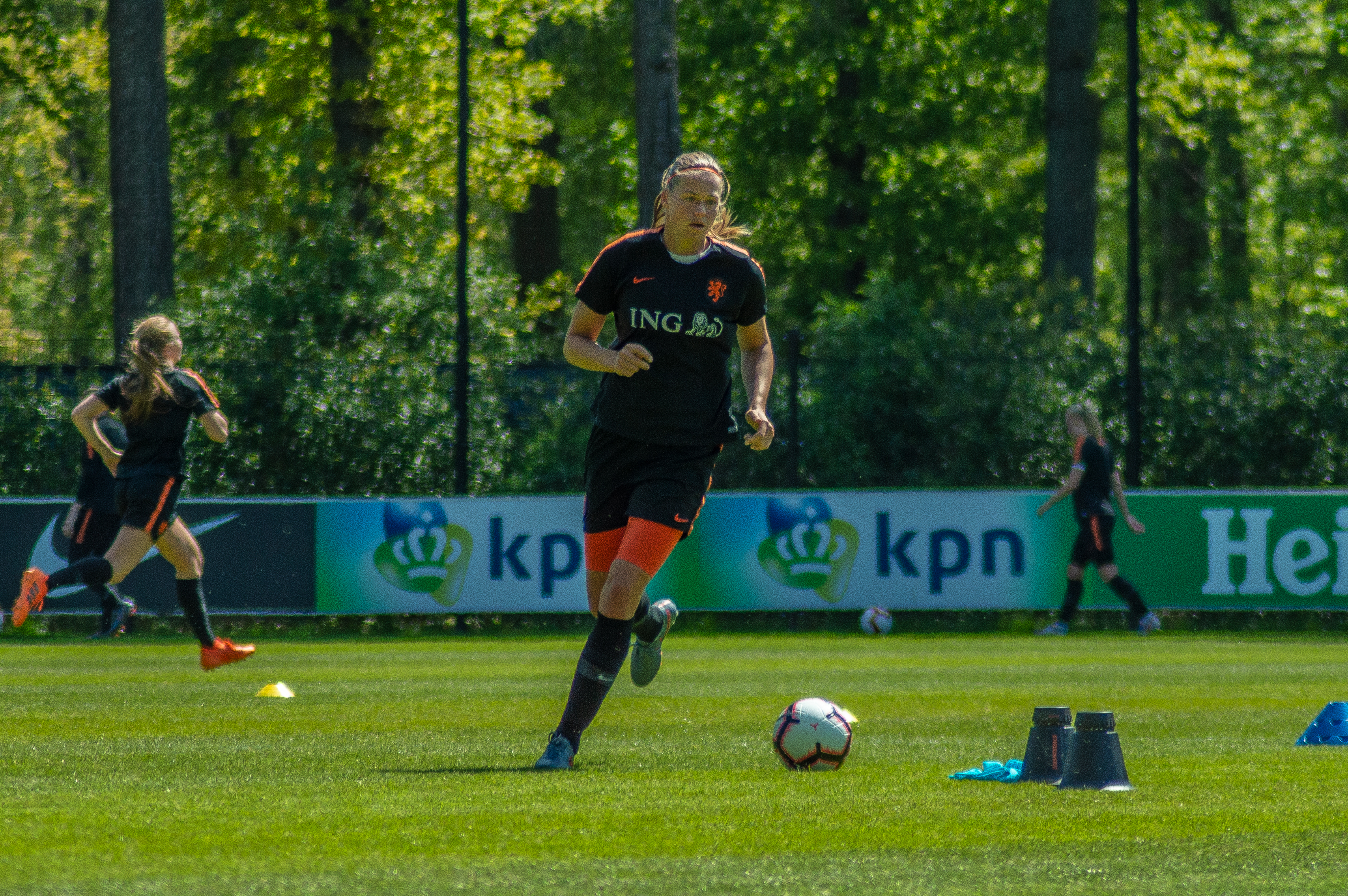 Eine Gruppe von Frauen, die Fußball auf einem grünen Feld umgeben von Bäumen und Pfosten spielt, mit einem Banner mit Text im Hintergrund, alle tragen Fußballtrikots.