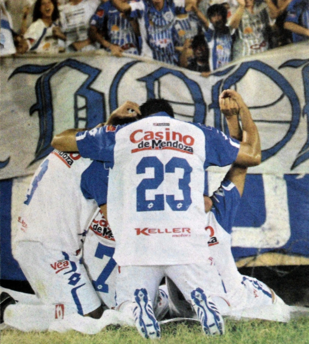 Gruppe von Fußballspielern, die auf dem Boden vor einer jubelnden Menge mit einer Fahne im Hintergrund feiern.