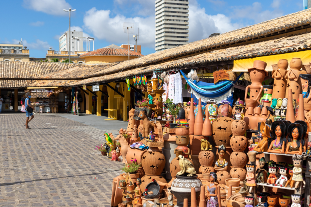 Ein belebter Markt in Cartagena, Kolumbien, mit verschiedenen Artikeln wie Puppen, Tägen, Pflanzen und Klamotten zum Verkauf, Menschen, die durch die Gänge schlendern, und Gebäude, Masten und einen bewälktem Himmel im Hintergrund.