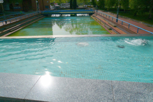 Großer rechteckiger Swimmingpool in einem Park, umgeben von Geländern und Laternenmasten, mit Bäumen und parkenden Fahrzeugen drumherum, vor Buildings und einem klaren blauen Himmel.