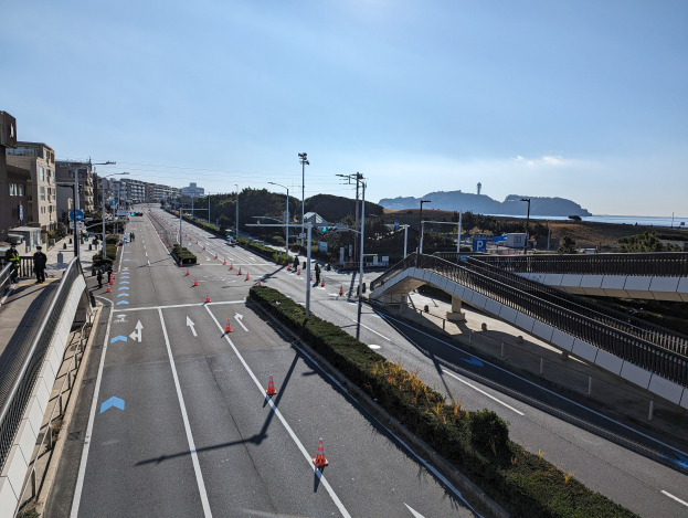 Straße mit Verkehrskegeln, Brücke mit Geländern, Laternenpfähle, Gebäude mit Fenstern und einige Menschen, vor Bergen und einem bewölkten Himmel, Teil des Takayama-Brücken-Projekts.