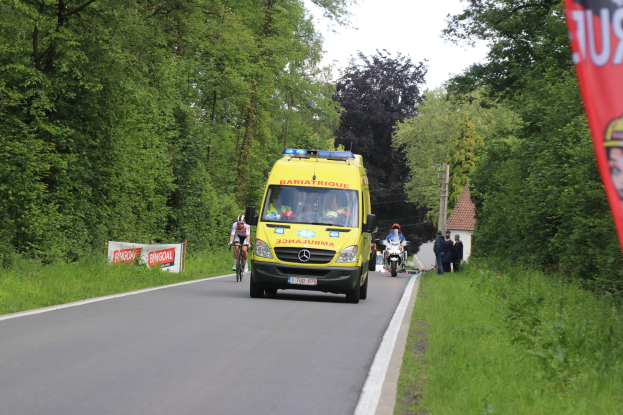 Ambulanz auf einer Straße mit Fahrradfahrern nebenher, Gras und Bäume auf beiden Seiten, Häuser und Masten im Hintergrund unter einem klaren blauen Himmel.