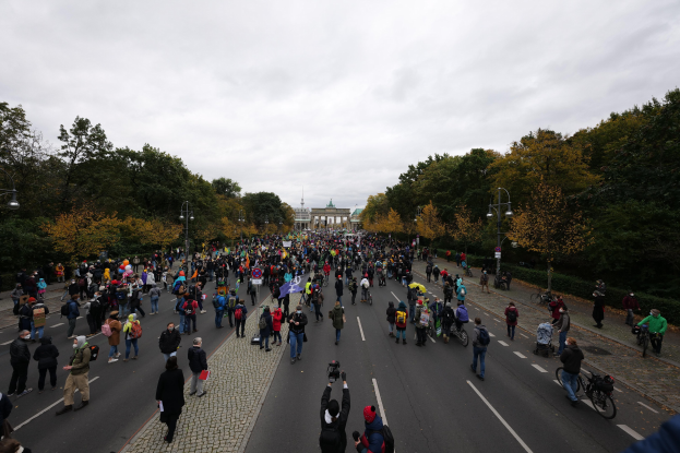Eine große Gruppe von Menschen marschiert auf einer baumbestandenen Straße mit Laternenmasten, hält Kameras, mit einem Gebäude und einem klaren Himmel im Hintergrund.