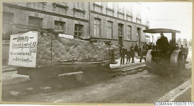 Schwarz-weiß-Foto eines Zuges auf den Schienen mit einer Gruppe von Menschen in der Nähe, einem Fahrzeug auf der rechten Seite und einem Gebäude mit Fenstern im Hintergrund.
