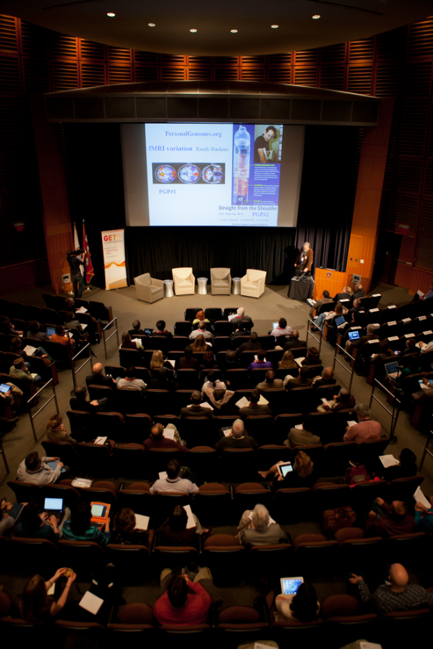 Ein mit sitzenden Menschen gefüllter Saal, der von vielen Lichtern beleuchtet wird, mit Fahnen auf der linken Seite, einem Podium auf der rechten Seite und einer Werbetafel auf der linken Seite.