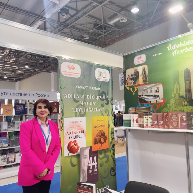 Eine Frau steht vor einem Stand auf einer Buchmesse in Moskau, umgeben von Büchern, Bannern, einem Stuhl und einem Tisch mit Büchern, unter einer Decke mit Lampen und Rohren.