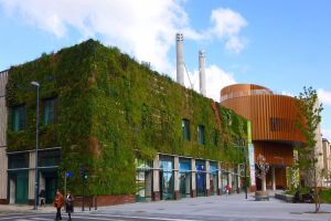Ein großes Gebäude mit einer grünen Wand an seiner Seite, umgeben von Straßeninfrastruktur, Fahrzeugen, Fußgängern, Bäumen und einem bewölkten Himmel, Teil des Green Building Project.