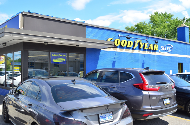 Außenansicht eines Goodyear Select-Autohauses mit parkenden Fahrzeugen, sichtbarer Beschilderung, Bäumen und einem bewölkten Himmel.
