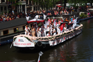 Eine Gruppe von Menschen auf einem Boot im Wasser, umgeben von Fahnen und Blumen, mit Gebäuden, Bäumen und einem Laternenmast im Hintergrund während der Amsterdam-Rot-Kreuz-Parade.