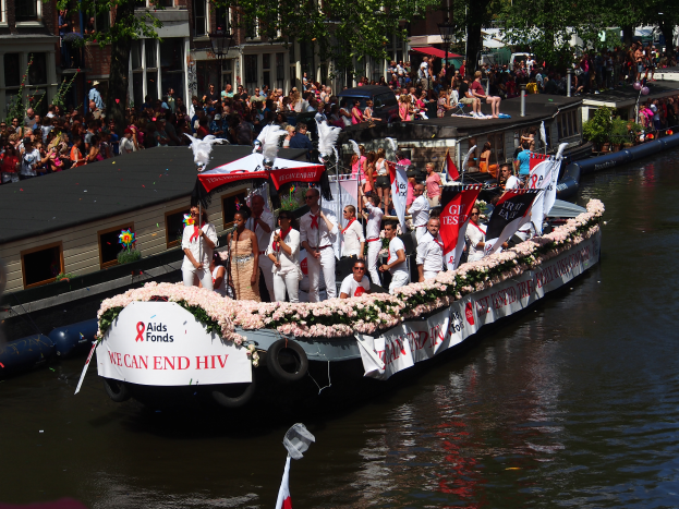 Eine Gruppe von Menschen auf einem Boot im Wasser, umgeben von Fahnen und Blumen, mit Gebäuden, Bäumen und einem Laternenmast im Hintergrund während der Amsterdam-Rot-Kreuz-Parade.