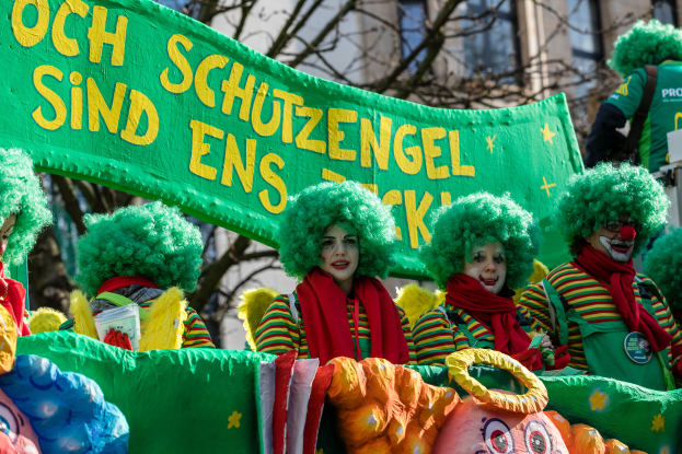 Eine Gruppe von Clowns in bunten Kostümen marschiert eine Straße entlang und hält ein Schild mit Text, mit Bäumen und einem Gebäude im Hintergrund.