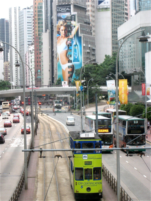 Eine belebte Stadtstraße mit Doppeldeckerbussen, Autos und anderen Fahrzeugen, umgeben von hohen Gebäuden, Bäumen, Polen, Laternen, Schildern, Bannern und einer Brücke unter einem klaren blauen Himmel, mit Text unten.