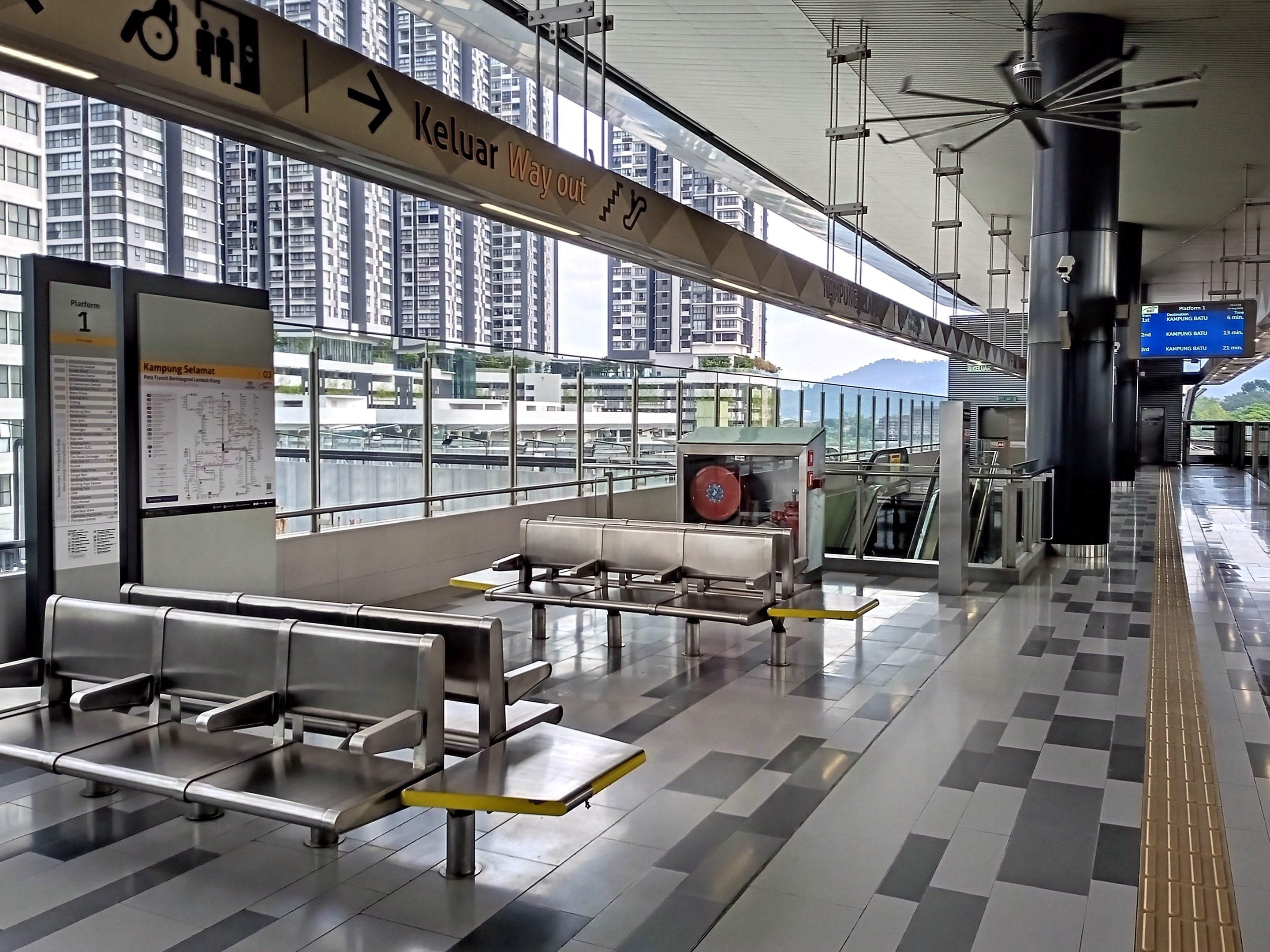 Leerer Flughafen Wartebereich mit mehreren Bänken, Informationsanzeigen, Säulen, Deckenventilator und Deckenbeleuchtung; Hintergrund zeigt Gebäude, Bäume, Wasser und einen klaren blauen Himmel.