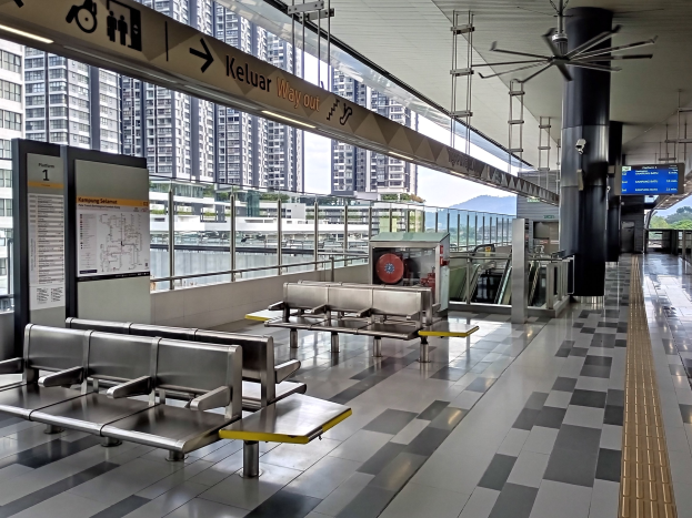 Leerer Flughafen Wartebereich mit mehreren Bänken, Informationsanzeigen, Säulen, Deckenventilator und Deckenbeleuchtung; Hintergrund zeigt Gebäude, Bäume, Wasser und einen klaren blauen Himmel.