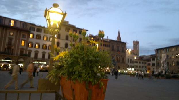 Eine Gruppe von Menschen, einige stehend und einige gehend, mit einer Pflanze, Statue, Lichtern und Gebäuden im Hintergrund.