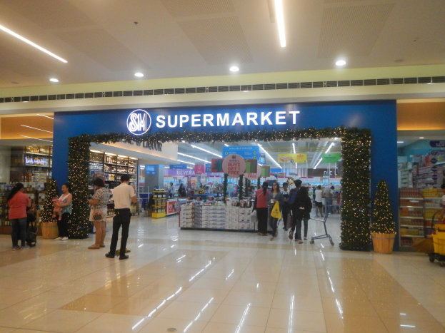 Eine Gruppe von Menschen steht vor einem Supermarkt, einige halten Taschen, mit einem Einkaufswagen, Topfpflanze, Regalauslagen, Schildern und Deckenleuchten.