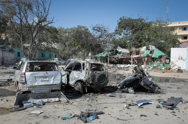 Die Auswirkungen eines Autobombenanschlags in Mogadischu, Somalia, mit verkohlten Überresten von Autos und anderen Gegenständen, die auf dem Boden verstreut sind, Bäumen, Gebäuden, Pfählen, Drähten und einer Wand im Hintergrund und einem sichtbaren Himmel.