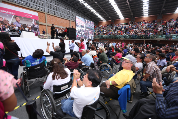 Eine große Gruppe von Menschen im Rollstuhl sitzt vor einem Publikum, mit Individuen auf der Bühne, die Kameras halten, einem Tisch mit einem Blumenstrauß und anderen Gegenständen, einem Banner mit Text und Bildern an der Wand dahinter und Lampen an der Decke.