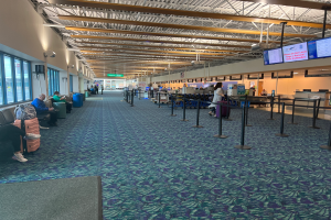 Eine lange Schlange von Menschen, die in einem Flughafen-Terminal warten, einige sitzen auf Stühlen, andere stehen, Stangen mit Bändern, Bildschirme an den Wänden, Lampen an der Decke und Glasfenster auf der linken Seite, Tafeln mit Text im Hintergrund.