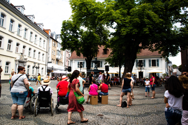 Eine Gruppe von Menschen, darunter einige im Rollstuhl, geht eine Kopfsteinpflasterstraße in Heidelbergs Altstad runter, mit Bäumen, Gebäuden, Laternenmästen und einer Statue im Hintergrund bei einem bewölktem Himmel.