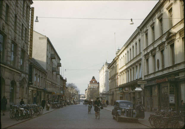 Altes Schwarz-Weiß-Foto einer Stadtstraße mit Menschen auf Fahrrädern und Autos, Gebäuden mit Fenstern auf beiden Seiten, parkenden Fahrrädern an den Seiten, Bäumen im Hintergrund und einem klaren blauen Himmel.