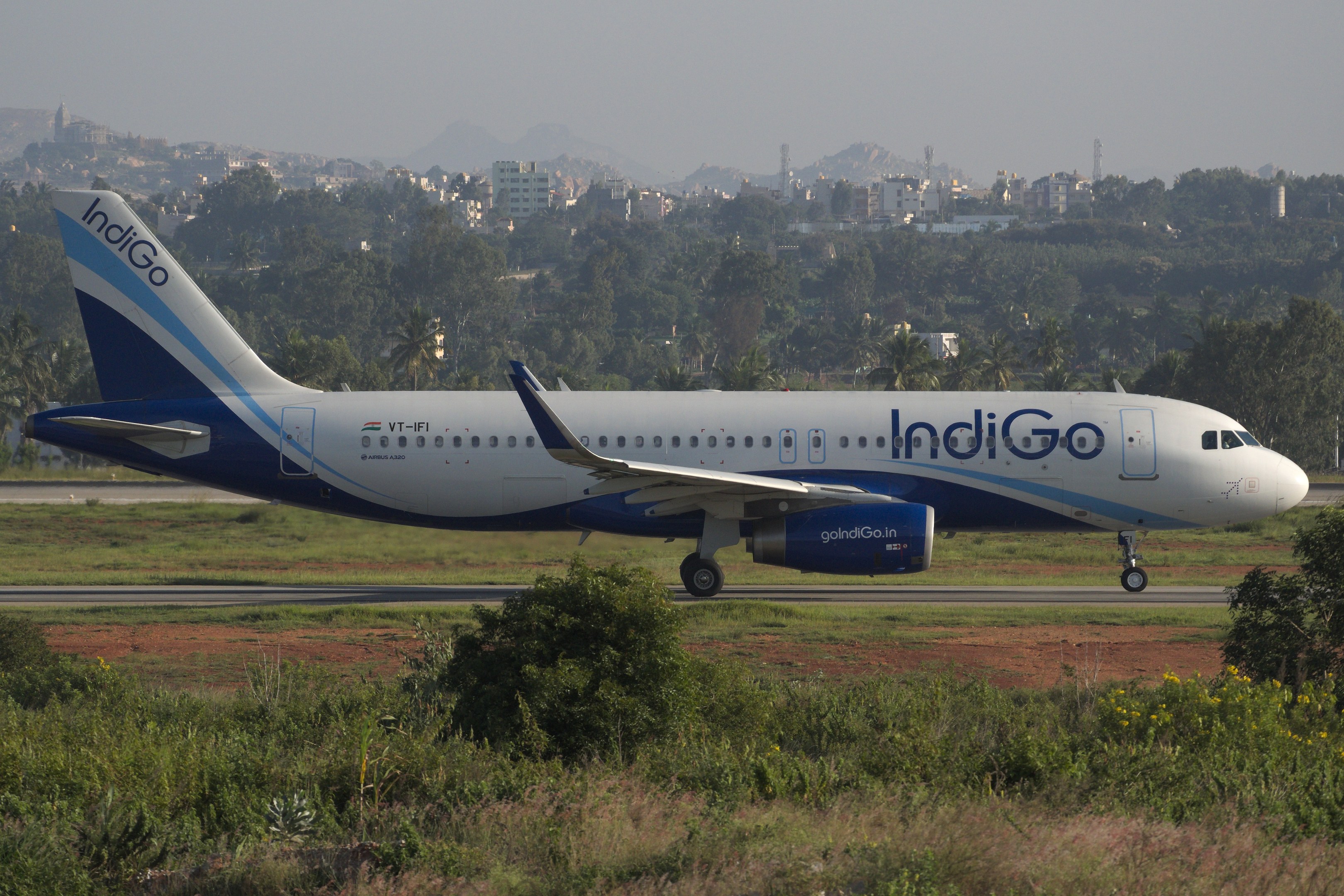 Indigo Airlines Airbus A320-200 auf der Rollbahn am Mumbai Airport mit Gras, Geb├Ąuden, Türmen, Bergen und Himmel im Hintergrund.