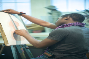 Ein Mann in einem Mantel und mit Uhr hält eine Zeichenunterlage, mit zwei Fenstern und einer Person mit Hut im Hintergrund.