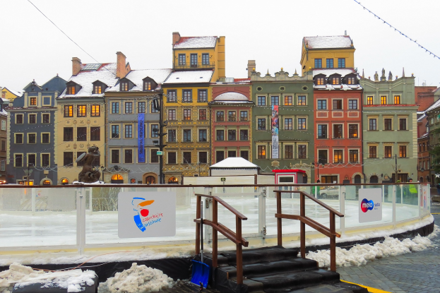 Eislaufbahn in einem Stadtplatz umgeben von Geb├Ąuden, Treppen, Gel├Ąndern, Informationsschildern, Laternen, B├Ąumen und schneebedeckter Erde unter einem Himmel.