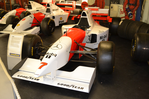 Rennwagen in einem Museum ausgestellt, mit Besuchern, die sie hinter einer Seilabsperrung beobachten, und informativem Text auf einer Tafel im Hintergrund.