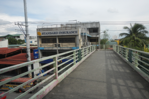 Eine Brücke mit Geländern, Fahrzeuge auf der Straße, Gebäude, Bäume, Strommasten mit Drähten und ein bewölkter Himmel im Hintergrund, mit einem Gebäude mit einer Tafel, auf der "Standard Insurance" steht.