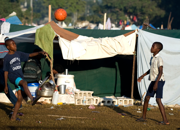 Eine Gruppe von Kindern spielt Fußball vor einem Zelt, das von Gras, Flaschen, Behältern und anderen Gegenständen umgeben ist. Im Hintergrund sind Bäume und ein klarer blauer Himmel zu sehen.