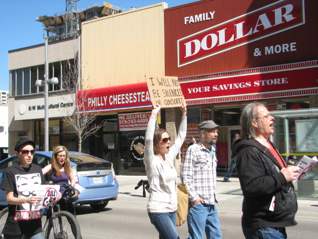 Eine Gruppe von Menschen demonstriert auf einer Straße mit Gebäuden, einem Pfahl und einem Baum im Hintergrund, einem Auto und einem Fahrrad auf der linken Seite und dem Himmel oben.