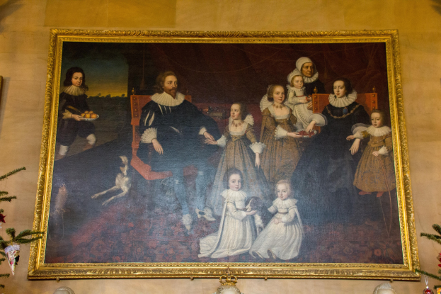 Gemälde einer Familie in einem Raum mit einem Weihnachtsbaum im Hintergrund, gerahmt und an einer Wand gehängt, mit zwei Foto Rahmen auf beiden Seiten und dekorativen Gegenständen unten.